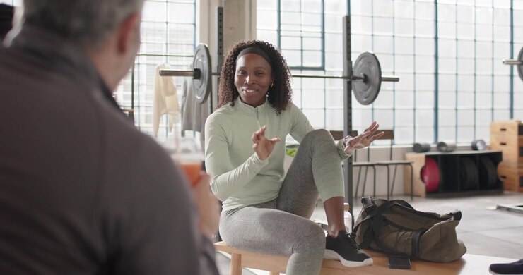 Diverse gym friends African American woman on bench listening to man speaking after set rubbing leg