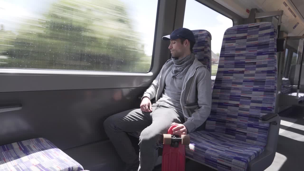 Young caucasian man seated in a train while holding a red mini skateboard in London city, England
