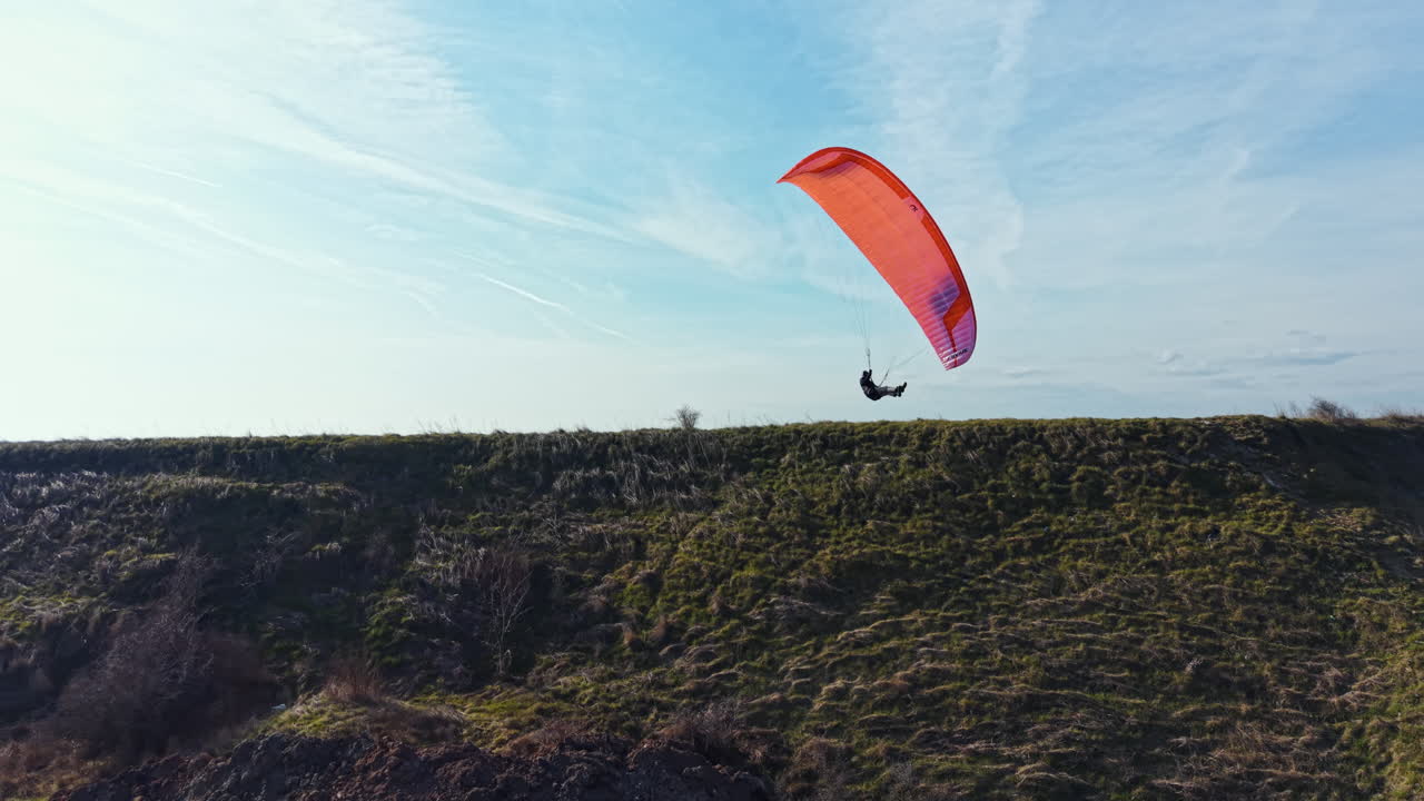 Parapente sobre una colina verde en un día despejado con cielo azul y pocas nubes de fondo, cerca de una zona rocosa en un espacio abierto.