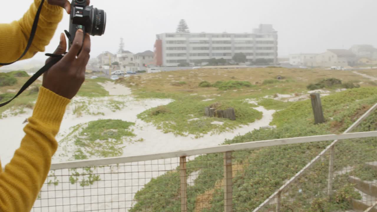 해변 4k에서 산책로에서 디지털 카메라를 가진 여성의 사진을 클릭하는 젊은 흑인 남자의 측면 보기