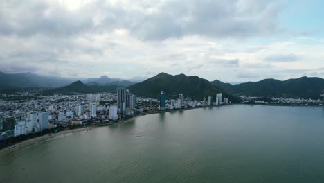 Luftdrohne,-Die-Während-Des-Sonnenuntergangs-Direkt-über-Die-Küste-Von-Nha-Trang-In-Der-Provinz-Khanh-Hoa-In-Vietnam-Schwenkt-Und-An-Einem-Bewölkten-Tag-Hohe-Gebäude-Und-Berge-Am-Horizont-Zeigt