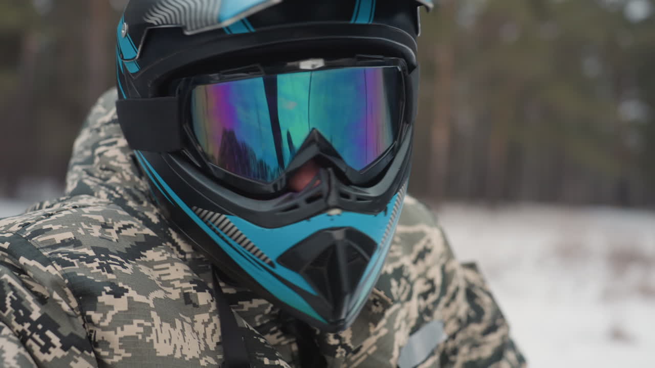 Side profile rider on snowmobile track grips handlebar and scans terrain, camouflage jacket and blue visor, ready posture, throttle poised, pine forest backdrop, cold breath and determined focus