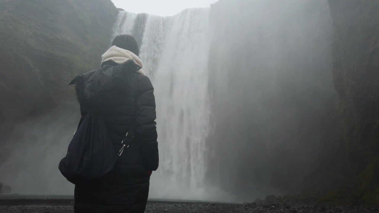 Rear view of a solitary female traveler admiring the powerful and scenic Skogafoss waterfall in Iceland. A moody and atmospheric landscape