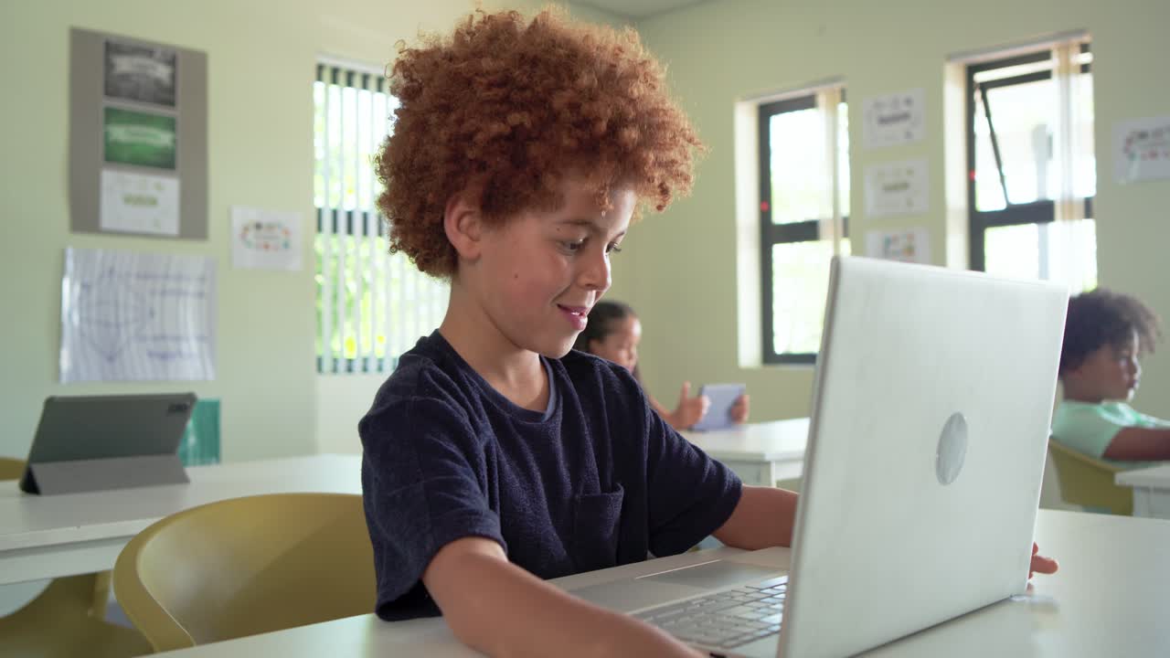 African American boy child typing, glancing, smiling on silver laptop in class while camera nearing