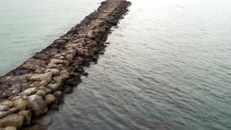 Drone-flyover-along-Jetty-rocks-with-beacon,-seagull-flying-above-water-surface,-Maritime-protection-Concept