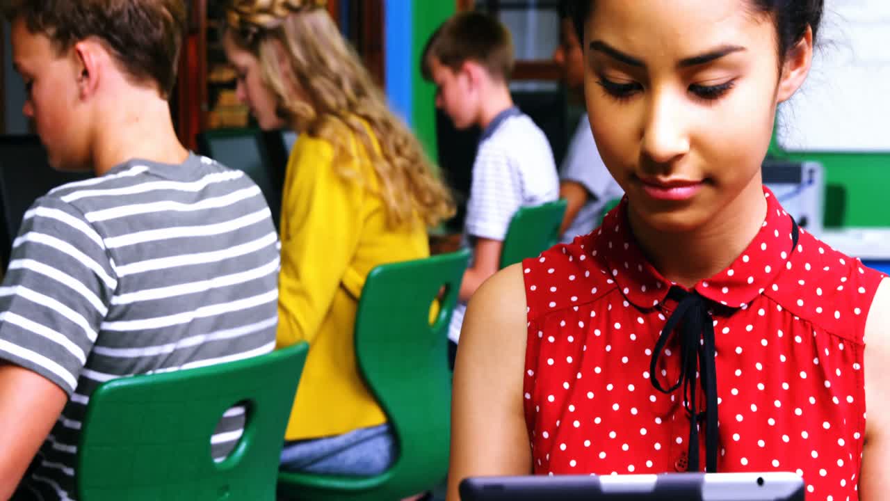 Schoolgirl using digital tablet while other students studying on computer in classroom