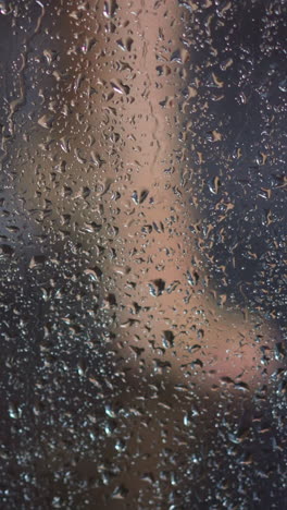 young woman washes feet in shower behind transparent fogged booth glass with flowing drops of warm water daily routine slow motion closeup