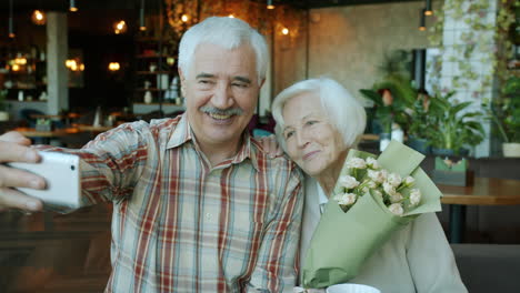 happy senior couple taking selfie with flowers in cafe
