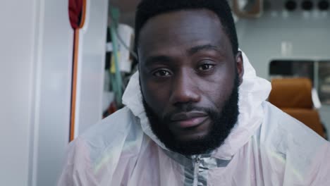 black paramedic in protective clothes looking at camera bearded african american doctor in hazmat suit blinking sleepily looking at camera while riding 911 service vehicle during pandemic