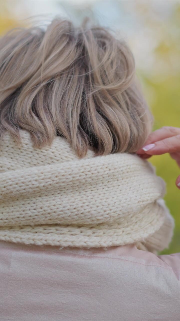 Primo piano della schiena di una donna con capelli biondi corti, che indossa una sciarpa lavorata a maglia color crema e un top rosa chiaro, su uno sfondo autunnale all'aperto. La scena si concentra sulla parte superiore del corpo e sui dettagli dell'abbigliamento