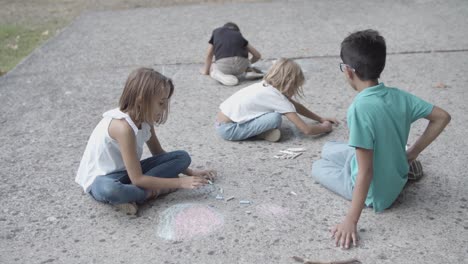 niños multiétnicos sentados sobre asfalto y dibujando con tizas
