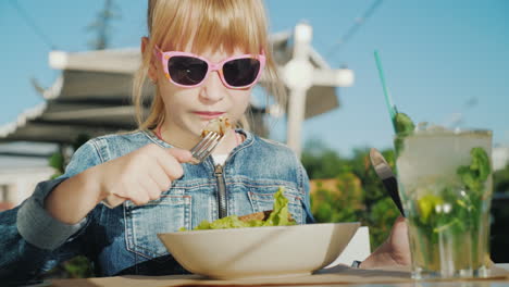 a girl in pink glasses is eating a salad on the summer playground of a cafe summer holiday with kids