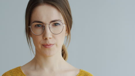 Retrato-De-La-Hermosa-Mujer-Con-Gafas-Y-Cabello-Largo-Y-Rubio-Mirando-Directamente-A-La-Cámara-Seriamente-Y-Luego-Sonriendo-Alegremente-En-El-Fondo-De-La-Pared-Blanca