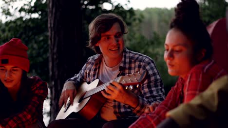 el guapo joven turista está tocando la guitarra y sonriendo mientras sus amigos están cantando y divirtiéndose descansando alrededor del fuego cerca del lago o el río. los árboles verdes son visibles.