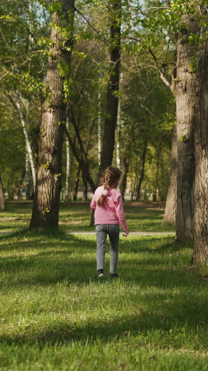 Joyful little girl jumps and runs along lawn in spring city park backside view. Happy child plays near old trees. Walking on sunny day