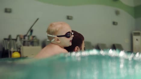 A-young-father-with-a-small-son-on-his-back-rowing-over-the-water.-Indoor-pool.-Camera-half-in-water.-Happy-child-with-father.-Slow-motion