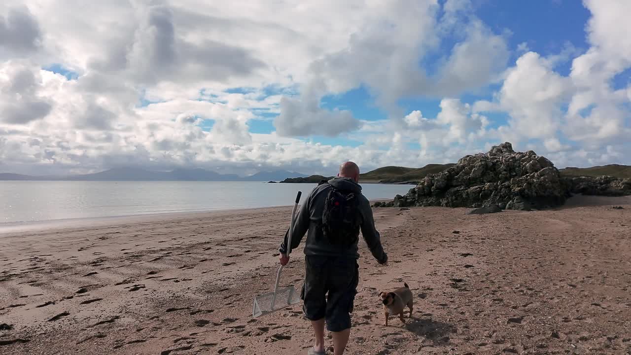 Bald male carrying sand scoop walking dog looking out across Welsh beach active lifestyle exploring