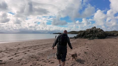bald male carrying sand scoop walking dog looking out across welsh beach active lifestyle exploring
