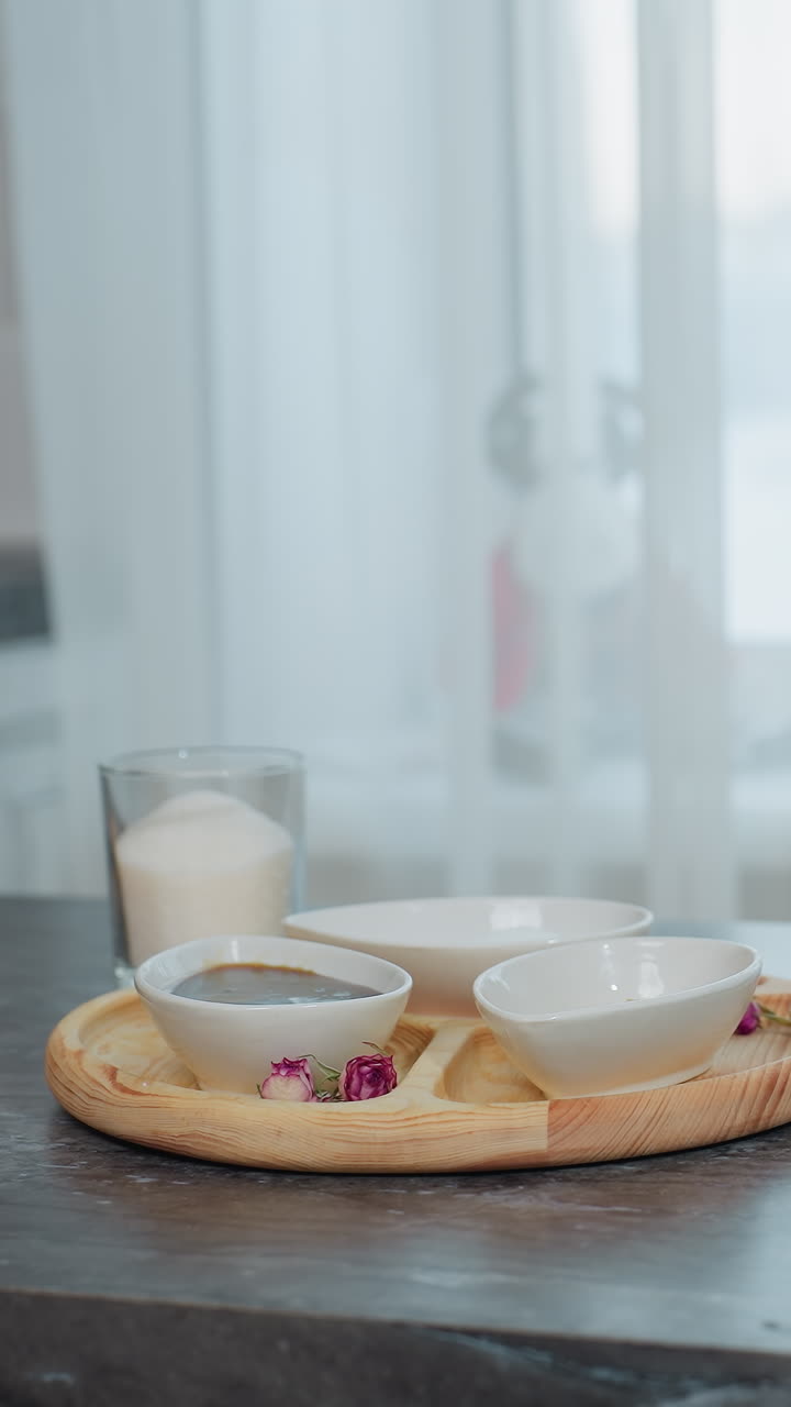 elegante mesa de cocina negra con bandeja de madera que contiene cuencos, especias, azúcar y mantequilla, rodillo colocado al lado del libro de portada marrón, cortina transparente blanca revela la vista del edificio a través de la ventana