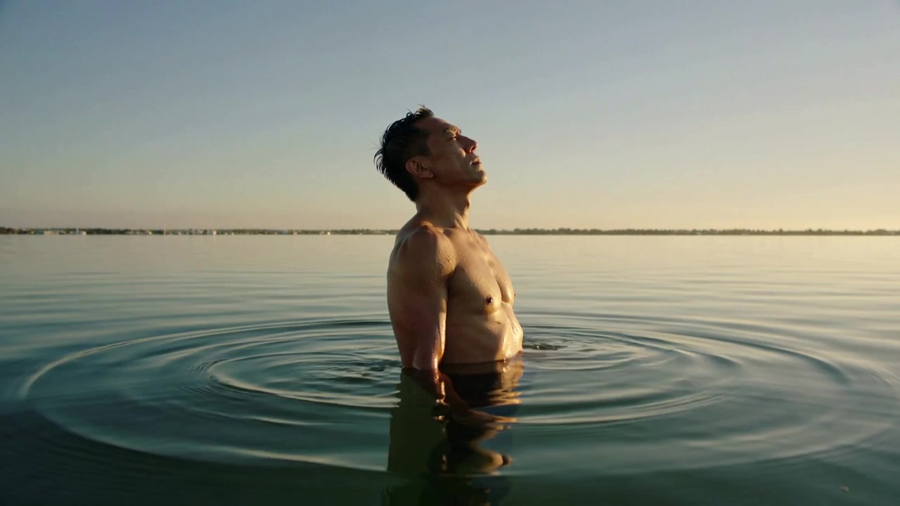 Hombre sin camiseta, en bañador oscuro, inhalando mientras se eleva e inclina hacia atrás para mirar el cielo en un lago, creando ondas.