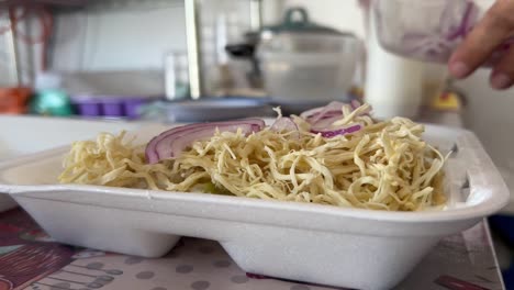 person adds sliced onions to shredded cabbage in a takeout container with blue tongs, close-up