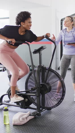 vertical video of diverse female fitness trainer and woman on exercise bike working out at gym