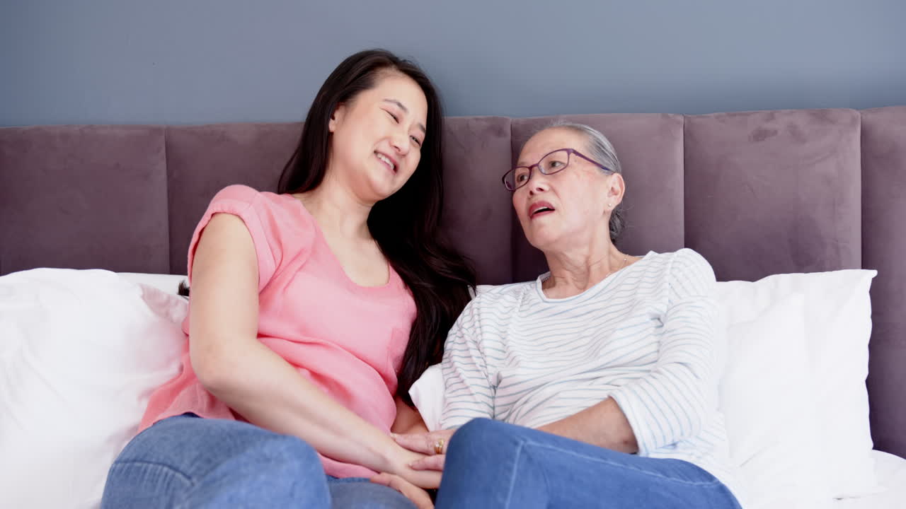 Sitting on bed, Asian grandmother holding hands with granddaughter, smiling together