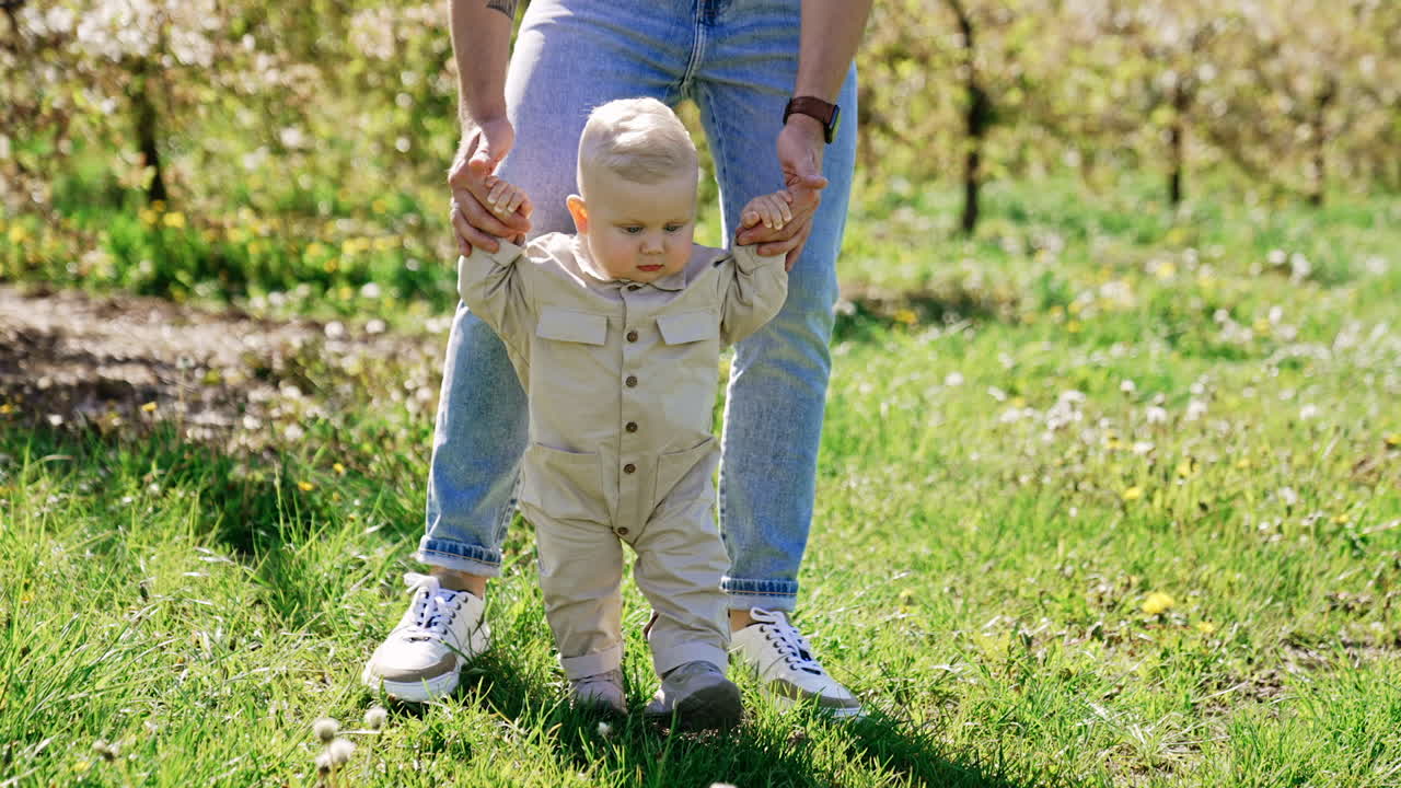 Unrecognized man wearing jeans leading a cute baby boy. Little child in a romper taking his first steps learning to walk. Nature at backdrop.