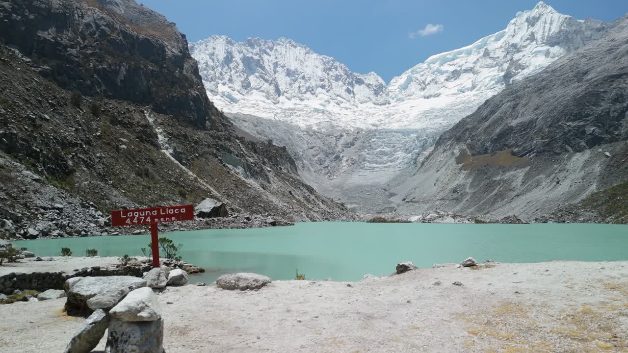 A first-person point-of-view shot shows a tourist hiking past the stunning, turquoise glacial waters of Laguna Llaca at the base of snow-capped peaks in Peru's Huascarán National Park