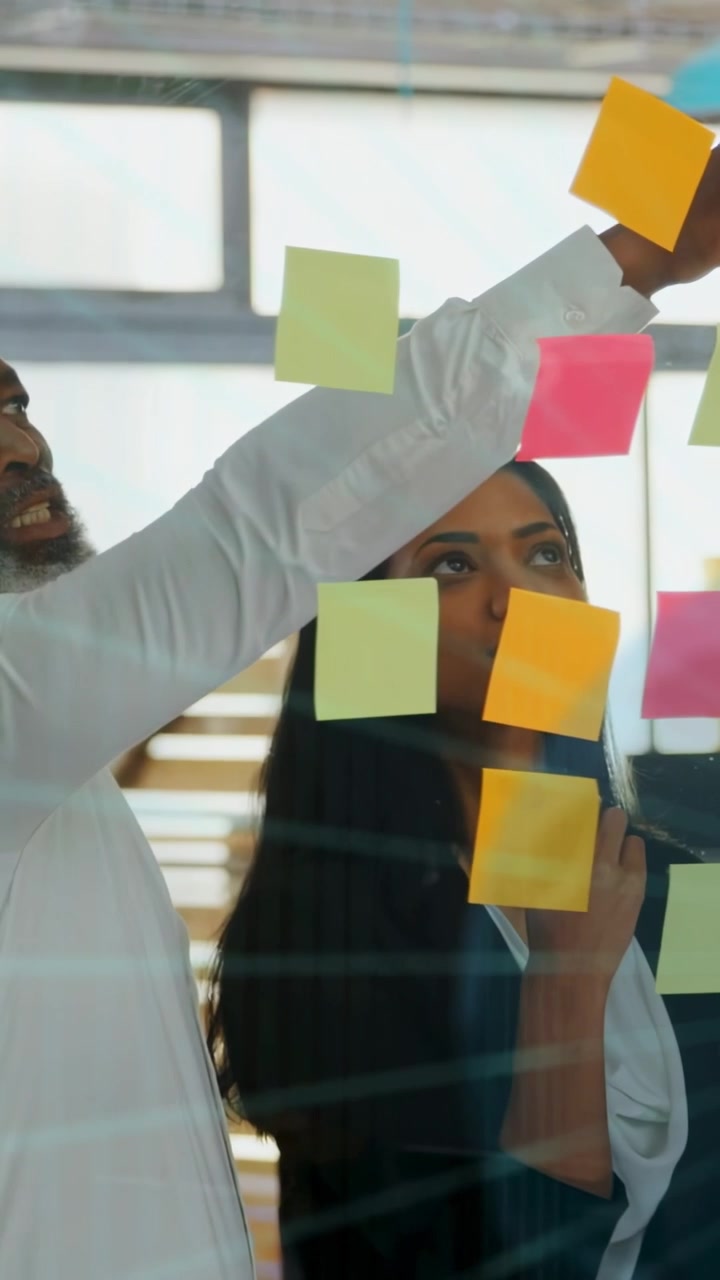 Vídeo vertical: Mão colocando nota verde, inspirando mulher em escritório de vidro, equipe organizando post-its brilhantes.