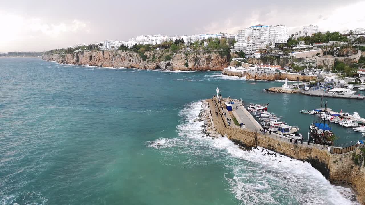 Drone view showing stone pier with lighthouse and boats beside cliffside town near Alanya harbor in Antalya region during windy weather with strong waves hitting rocky shoreline