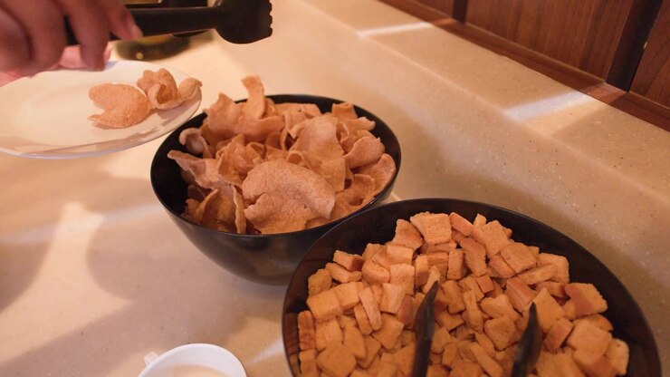 A person uses black tongs to serve crispy shrimp crackers from a bowl onto a plate.