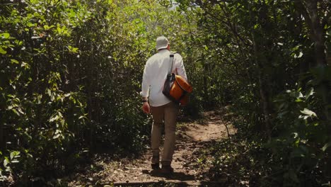 a man with a cap, a white shirt and a satchel walks through a mauritian jungle