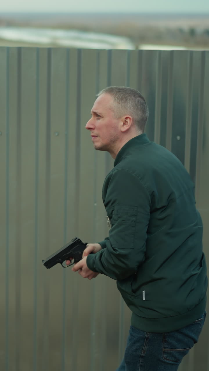 녹색 재을 입은 남자가 권총을 들고 산업 구역의 금속 울타리를 따라 조심스럽게 움직이고 있습니다.