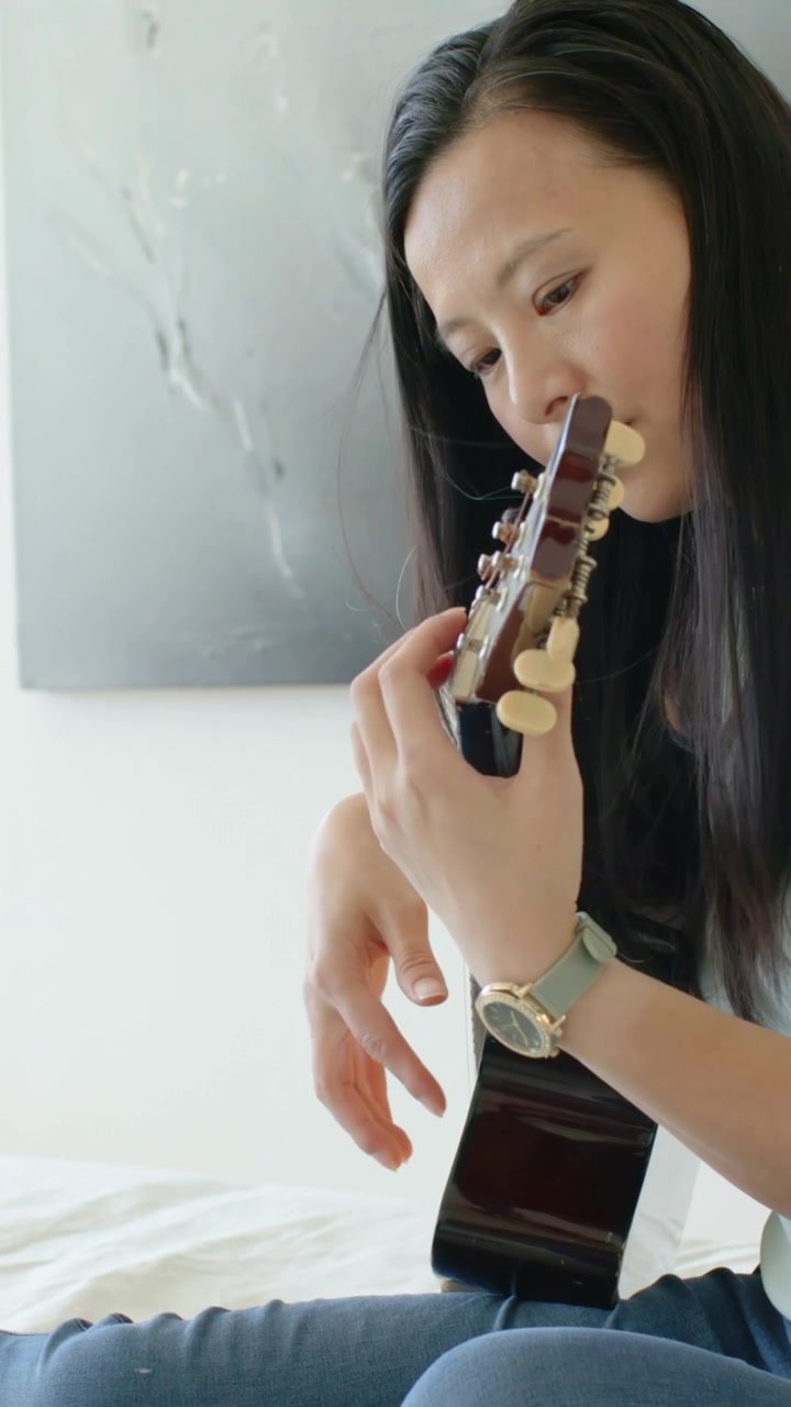 Woman tuning uke and playing strings on bed for music practice, camera panning right showing lamp