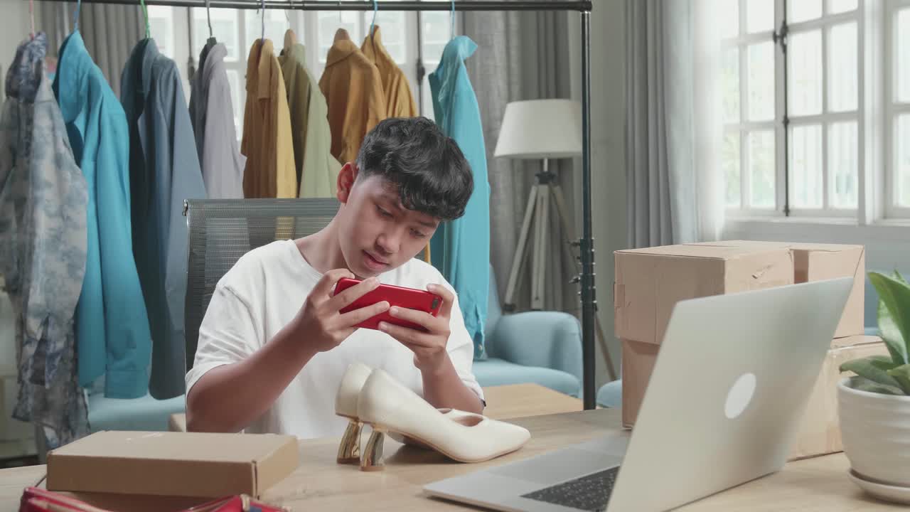 Asian Man Online Seller Taking Photos With Mobile Phone While Using Computer For Selling Clothes At Home