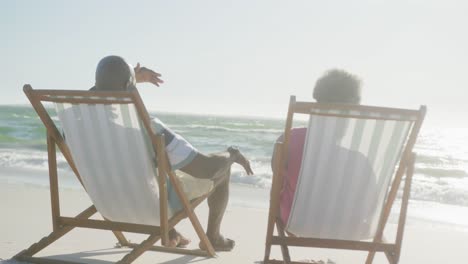 Happy-senior-african-american-couple-sitting-on-deck-chairs-at-beach,-in-slow-motion