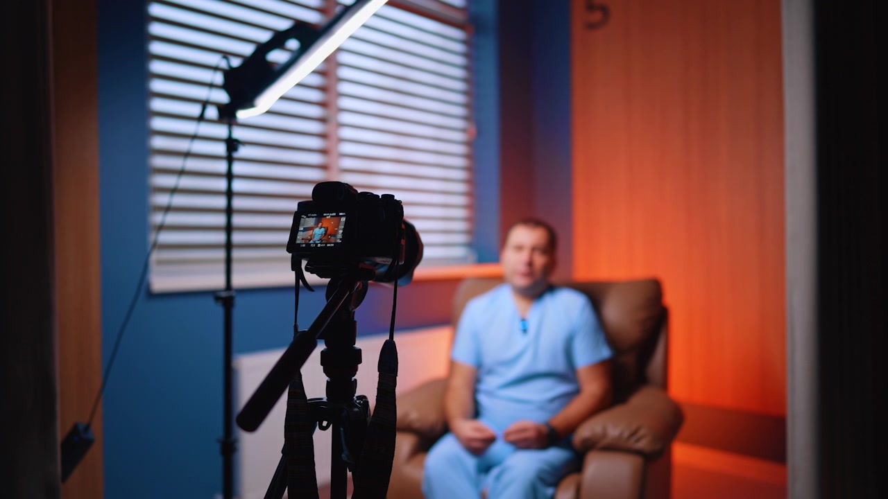 Câmera filmando homem sentado em poltrona com detalhe de parede laranja. Rig e boom em foco no primeiro plano, apresentador desfocado de uniforme médico emoldurado contra tons azuis e laranjas.