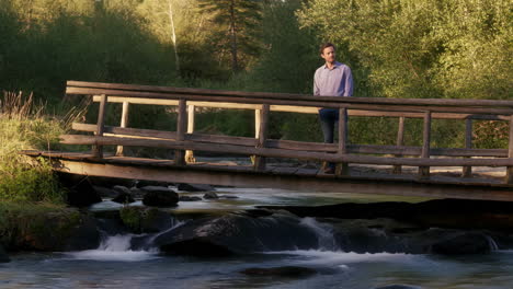 A man stands on a wooden bridge over a flowing river in a natural setting