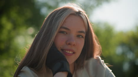 close-up of young woman leaning on bike handlebar, hand resting on chin with a gentle, contemplative smile, sunlight softly illuminates her face against a blurred background of lush green foliage