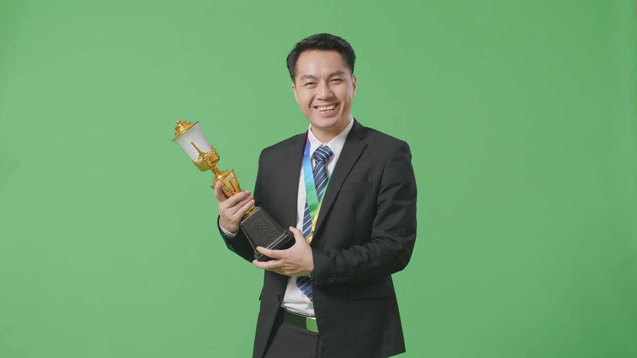 슈트와 타이를 입고 금메달을  아시아 사업가, 손에 금 트로피를 들고 스튜디오의 녹색 스크린 배경에서 첫 번째 우승자로 카메라에 미소 짓고 있습니다.