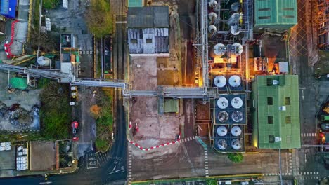 forward moving aerial footage of a large industrial plant at dusk, showing pipework structures, buildings, cooling towers, steam, and work vehicles