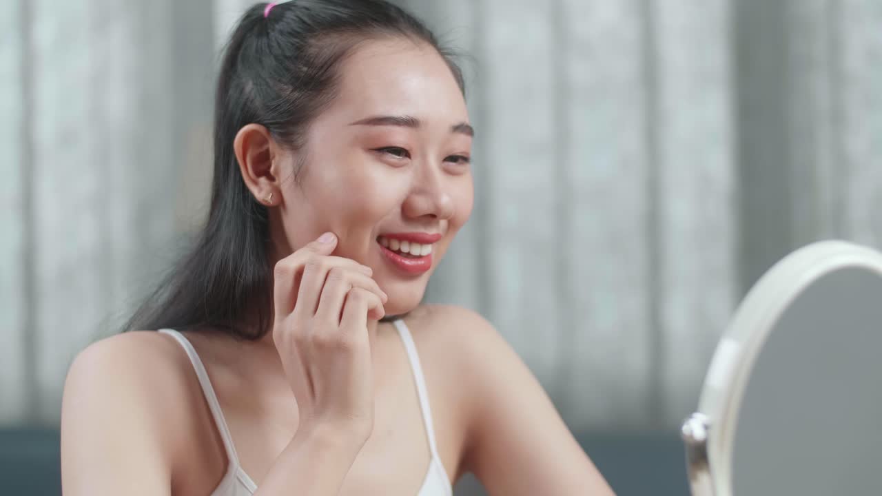 아름다운 젊은 아시아 여성이 거울을 바라보고 집에서 메이크업을 한 후 미소 짓고 있습니다.