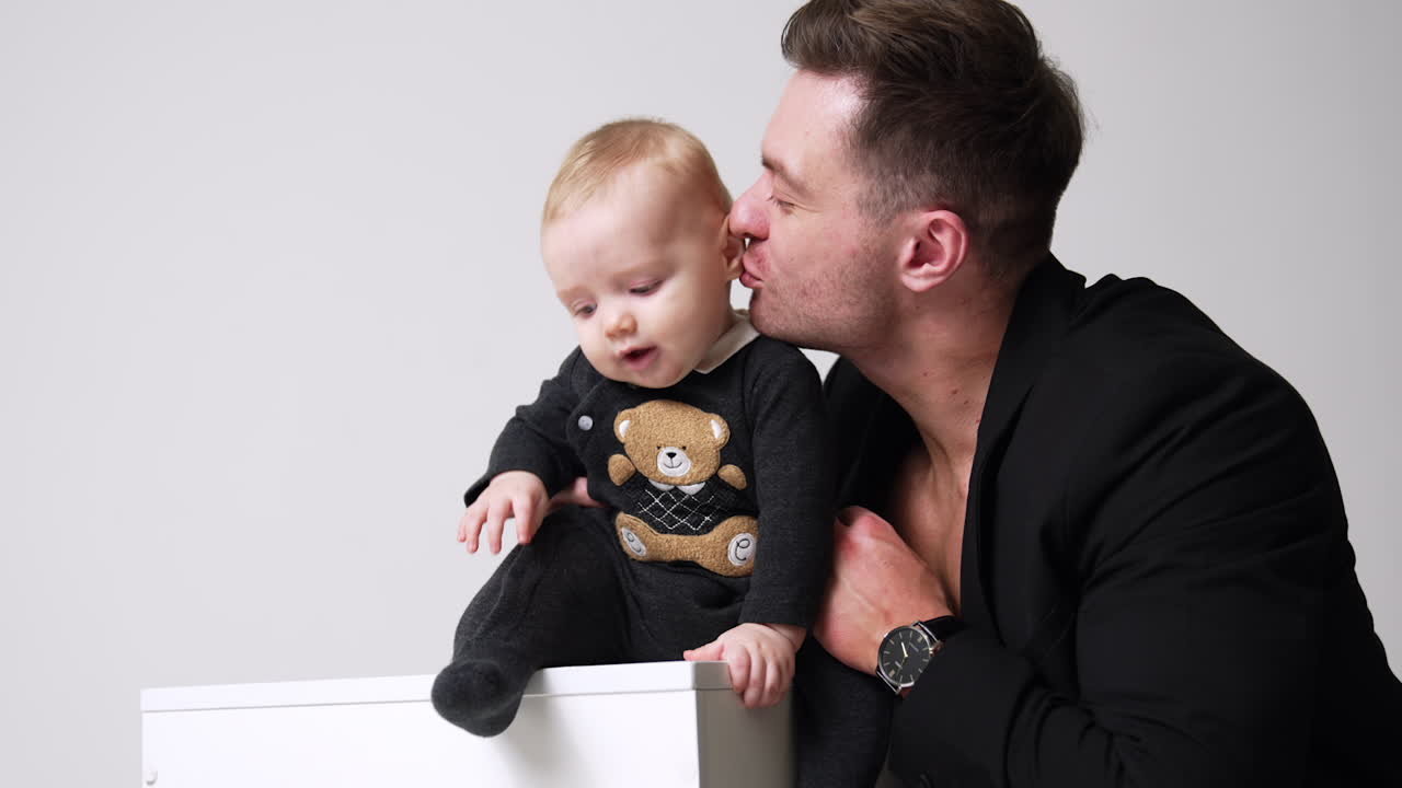 Adorable smiling baby sitting on the cube. Father holding his son and kissing him lovingly. White backdrop.