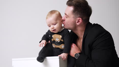 adorable smiling baby sitting on the cube. father holding his son and kissing him lovingly. white backdrop.