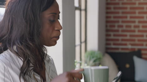 portrait-happy-african-american-woman-using-smartphone-drinking-coffee-at-home-browsing-social-media-messages-enjoying-relaxed-morning-looking-out-window-texting-on-mobile-phone