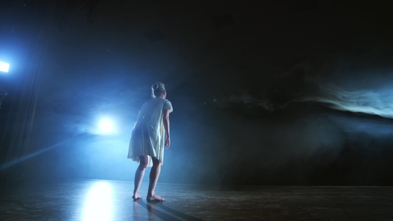 una joven bailarina baja descalza al escenario y se mueve en cámara lenta en un vestido blanco suelto.