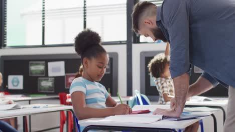 diverse male teacher and happy schoolchildren sitting at desk in school classroom