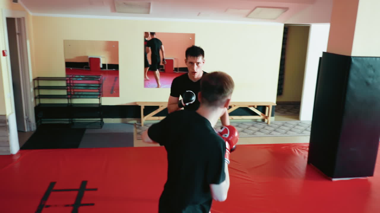Karate practitioners engaged in sparring session on red mat inside martial arts gym, fighter lifts knee striking while opponent stays guarded, showing combat focus and determination