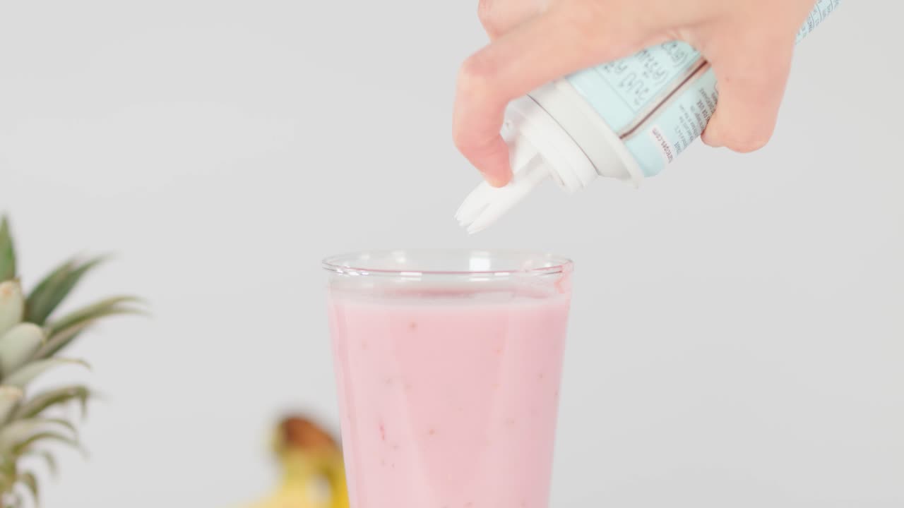 Hand dispenses whipped cream onto pink smoothie in glass, bright studio lighting, fruit background visible